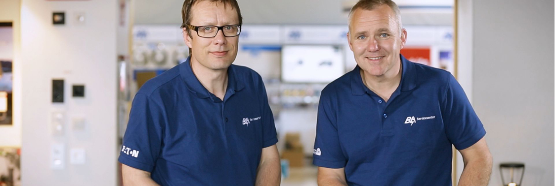 To BA-ansatte smiler mot kamera fra innsiden av et servicesenter