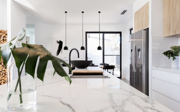 Stort, lyst og moderne kjøkken med kjøkkenbenk i hvit marmor, hvite underskap, overskap i lys eik og sorte detaljer rundt vinduer samt sorte lamper som henger over kjøkkenbenk.