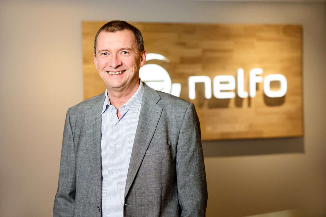 En mann med mørkt hår i grå blazer og hvit skjorte står foran en vegg med Nelfo-logoen.