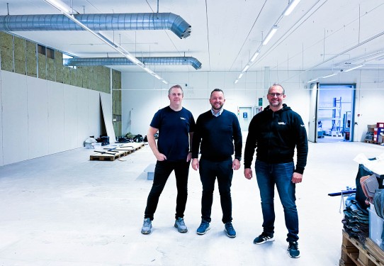 tre menn i blå arbeidsklær står ved siden av hverandre og smiler til kamera i tomt næringslokale.
